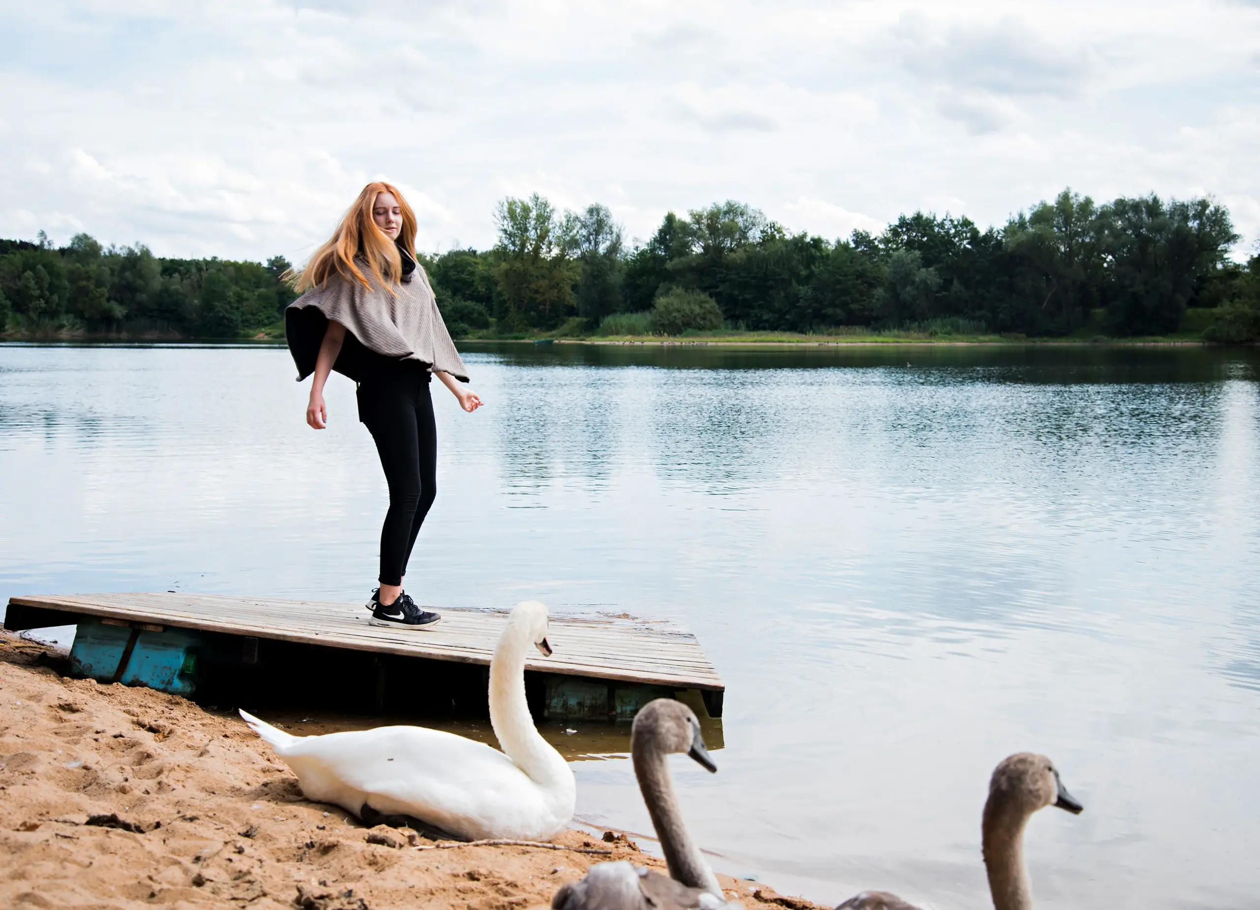 Junge Frau steht entspannt auf einem Holzsteg an einem ruhigen See, im Vordergrund ruhen Schwäne am Sandufer – Symbol für natürliche Ruhe und Ausgeglichenheit.